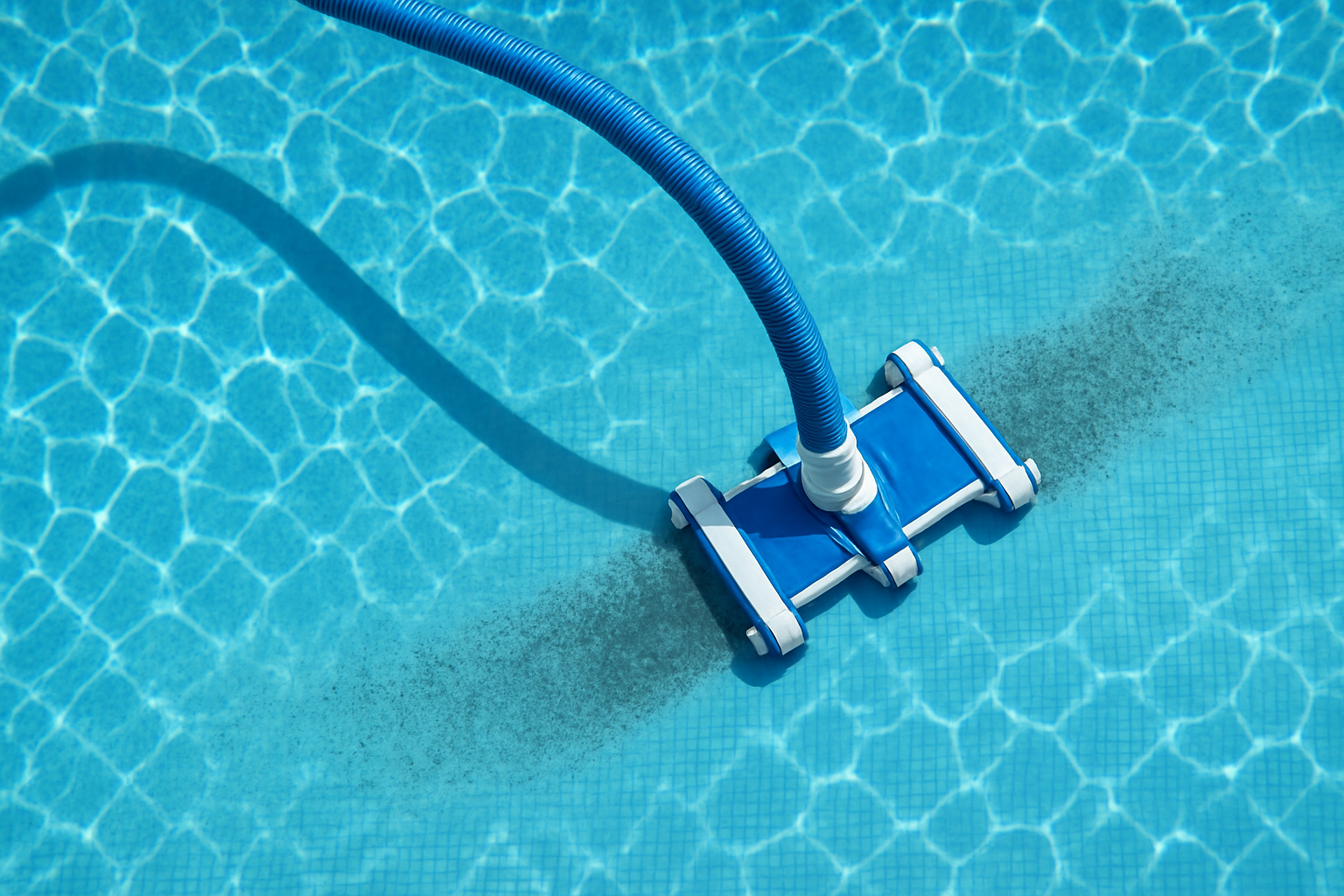 Top-down view of a pool vacuum cleaning equipment working underwater, showing the vacuum head on the pool floor with its hose extending across the crystal clear blue water, with sunlight reflections and visible contrast between cleaned and uncleaned areas